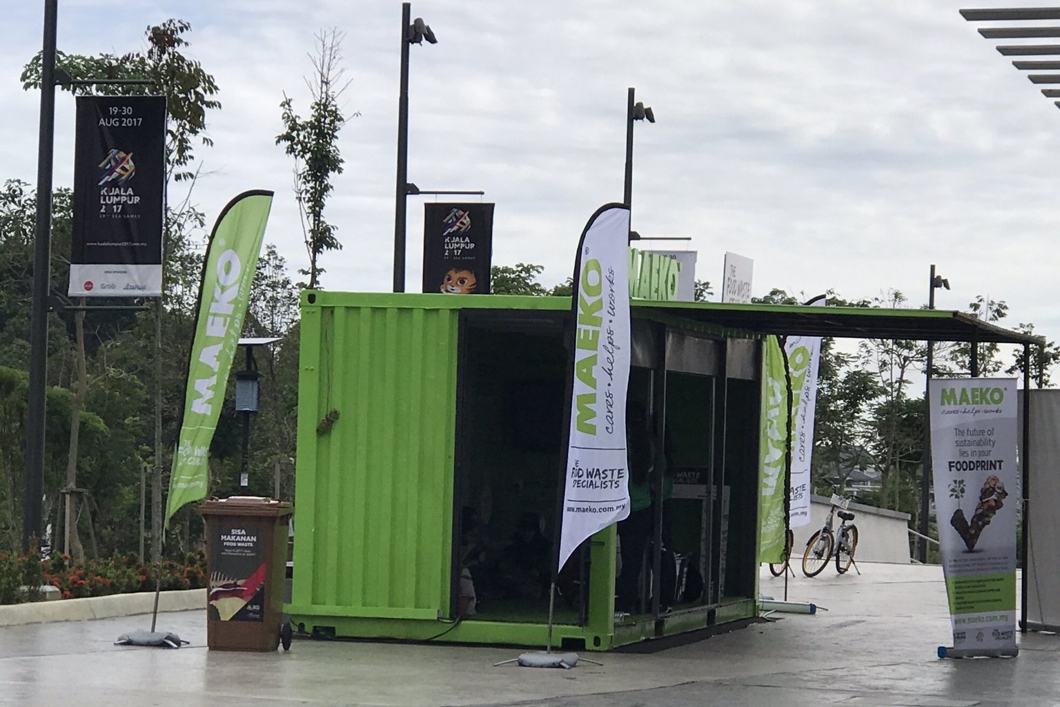 MAEKO’s bright green shipping-container composting unit, open along one side, standing on a paved public plaza with MAEKO flags around it.