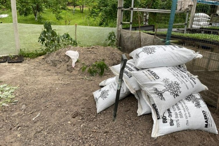 Sacks of MAEKO compost stacked on bare earth beside a planting bed on a farm.