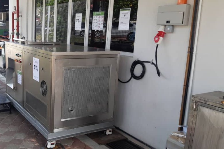 A MAEKO composter installed outside a factory canteen, its power cable run to a wall socket.