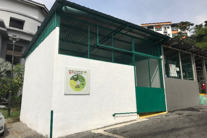 A white composter house with a green mesh roof and a MAEKO sign, built in a car park.