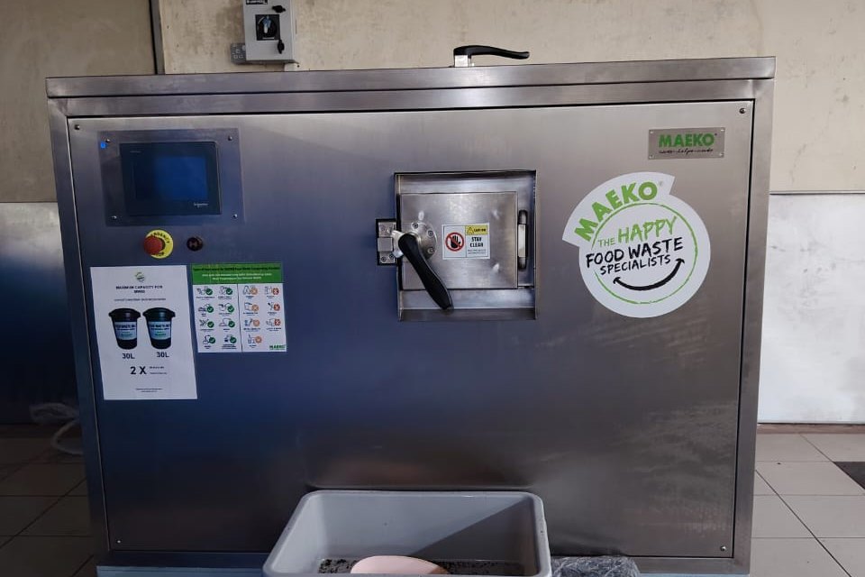 The front of a MAEKO composter with a touch screen, a feed hatch and a discharge tray, in a hospital kitchen.