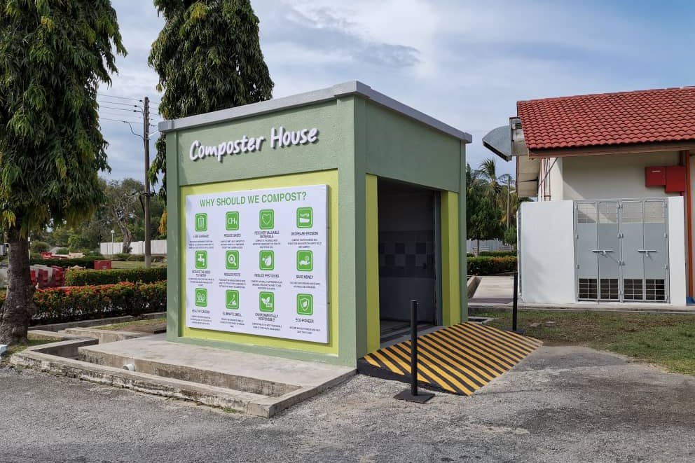 A small green-and-grey building signed Composter House on a campus lawn, with a board explaining why we compost.