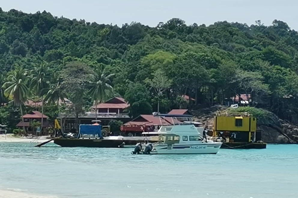 A boat landing on a beach below a forested island hillside, with the resort buildings along the shore.