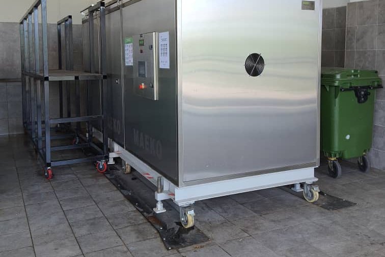 A stainless-steel MAEKO composter in a tiled service corridor, a green wheelie bin beside it.