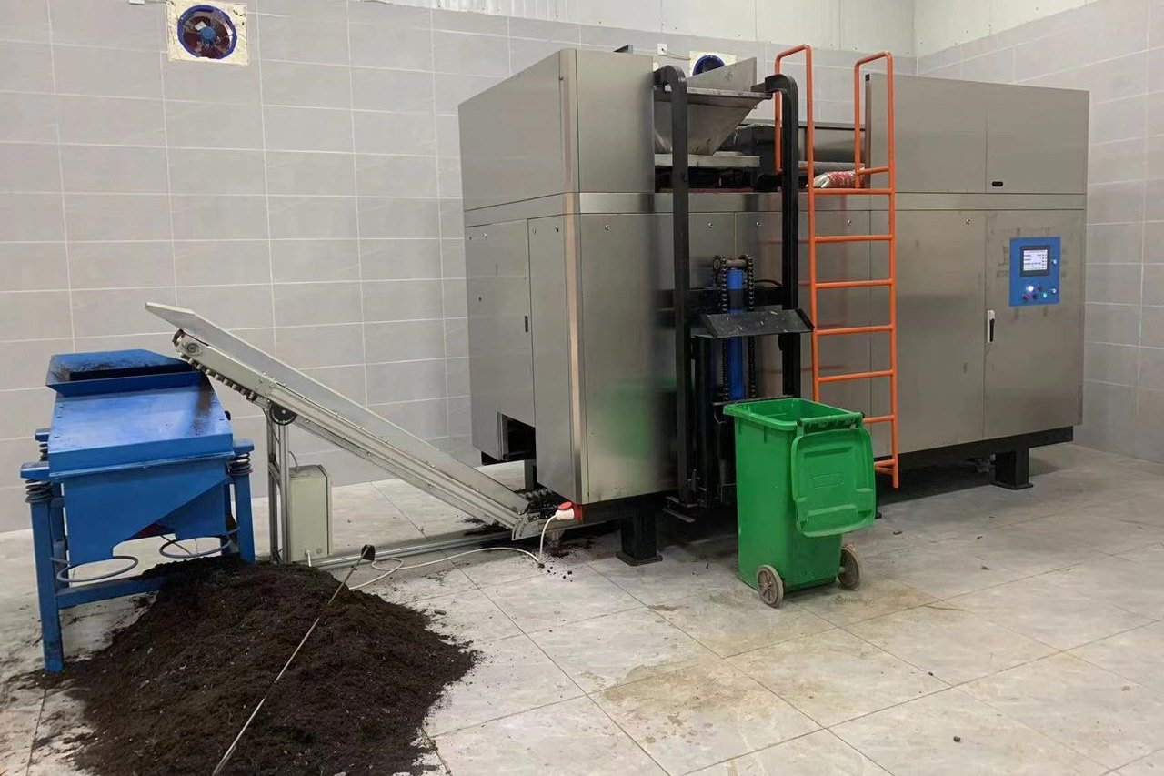 A large MAEKO composter with an orange access ladder in a tiled plant room, finished compost heaped below its discharge.
