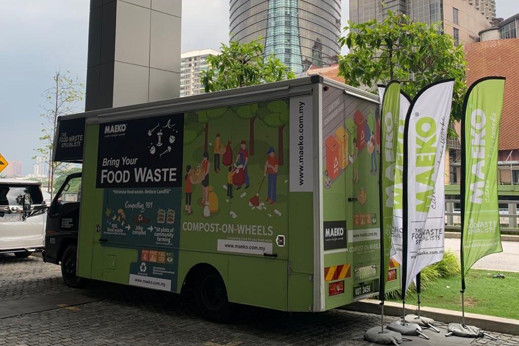MAEKO's green Composting-on-Wheels truck parked between office towers, its side painted with a garden scene and the words Bring your food waste, with three MAEKO flags beside it.