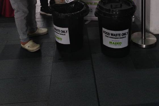 Two black bins labelled Food waste only at a MAEKO stand, a hand tipping scraps into the first one.