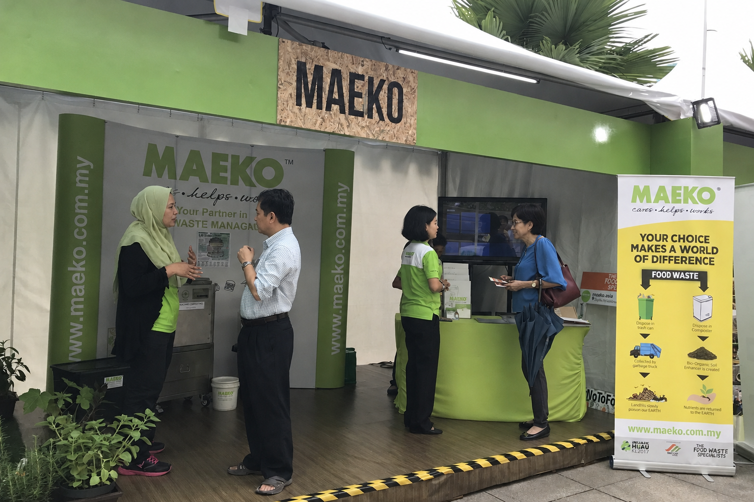 MAEKO’s stand at an event: staff explaining a small stainless composter to visitors across a counter of leaflets, under a MAEKO backdrop.