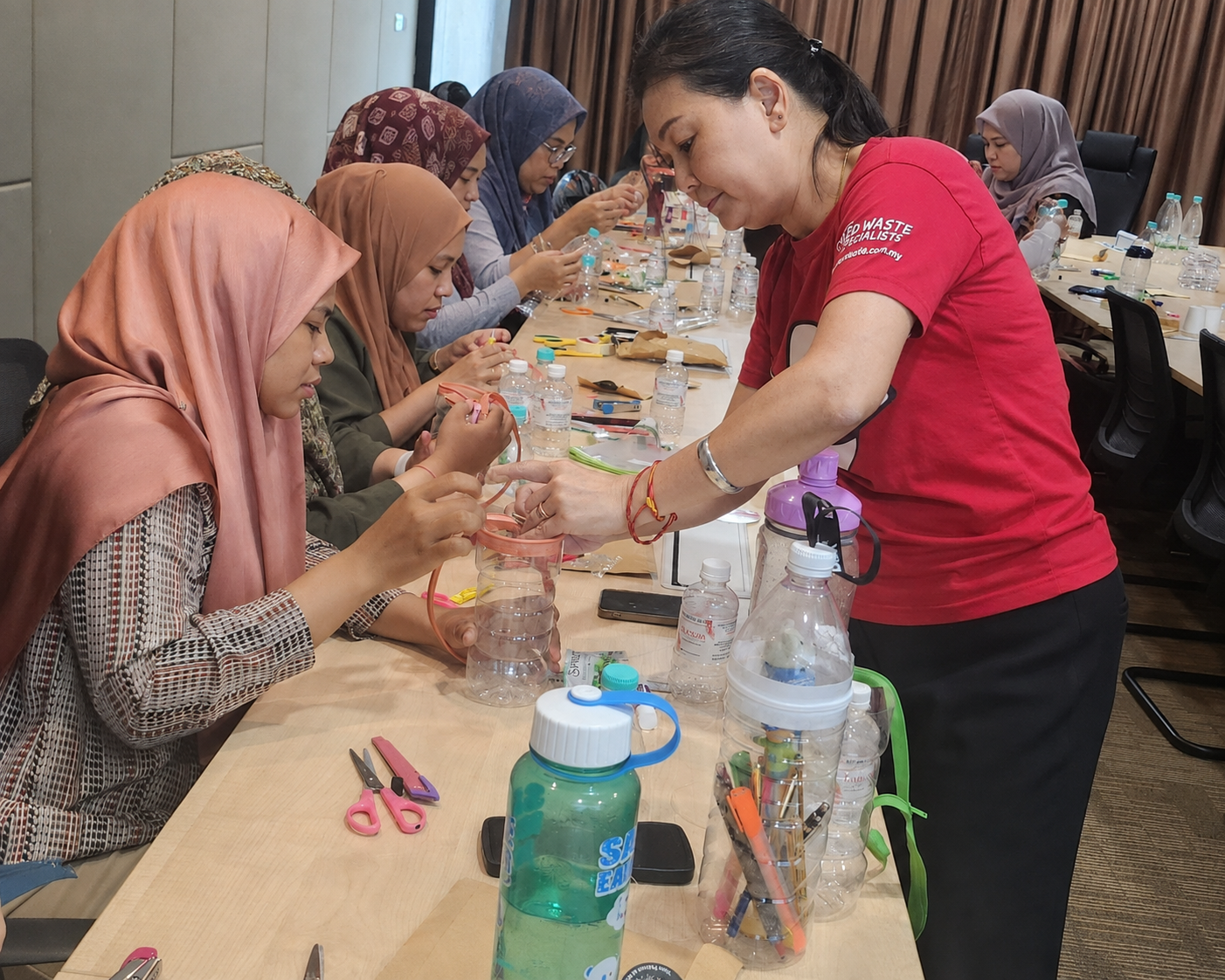 A MAEKO trainer helps a seated participant fit a handle to a cut-down plastic bottle, with a row of adults doing the same along the table.